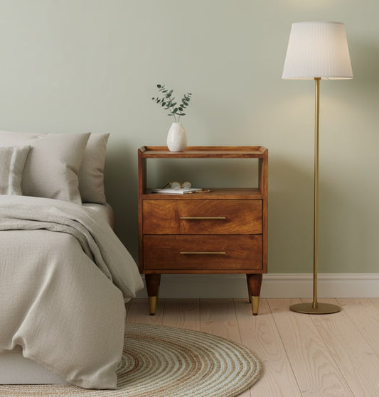 A wooden bedside table with simple design, featuring two drawers and a small shelf above. It's placed next to a bed with white bedding and a light floor lamp in a softly lit room.,