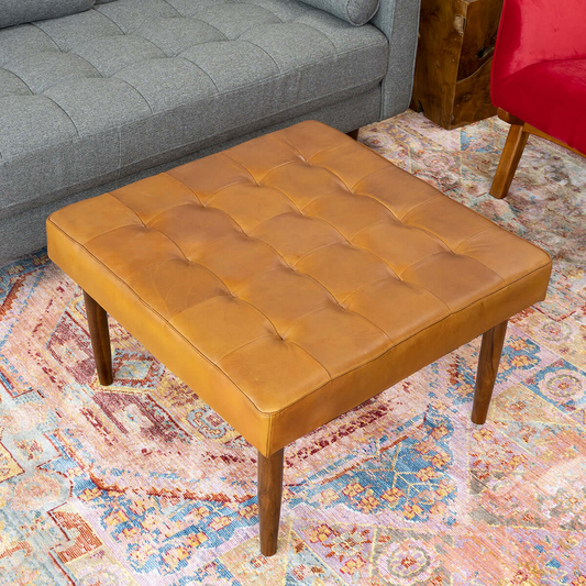Tufted square leather ottoman with wooden legs on patterned rug, gray couch in background