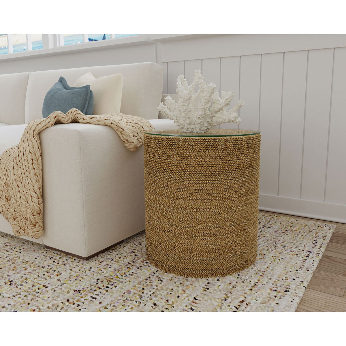 A light brown woven side table with a transparent glass top, sitting on a speckled rug beside a beige sofa with pillows and a beige throw, in a minimalistic interior setting.