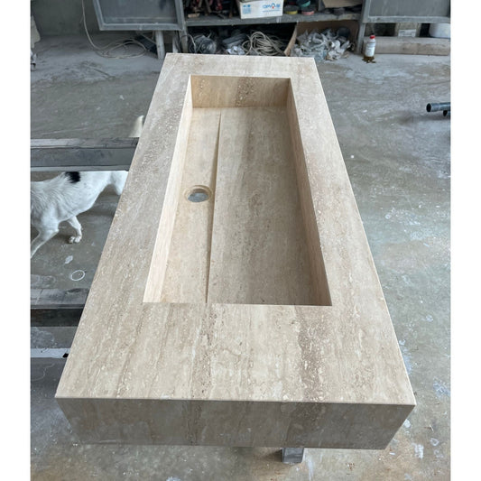 Rectangular travertine sink with a natural beige color and veined texture on an industrial concrete floor with a dog in the background, showing its handmade quality without artificial finish.