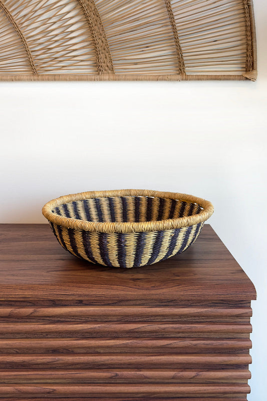 Bolga tray made of natural materials with black stripes placed on a wooden shelf against a minimalist wall hanging background.