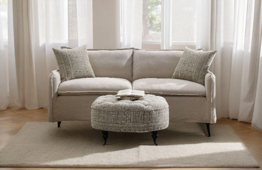 Beige linen sofa with patterned pillows and matching ottoman on a white rug in a sunlit room