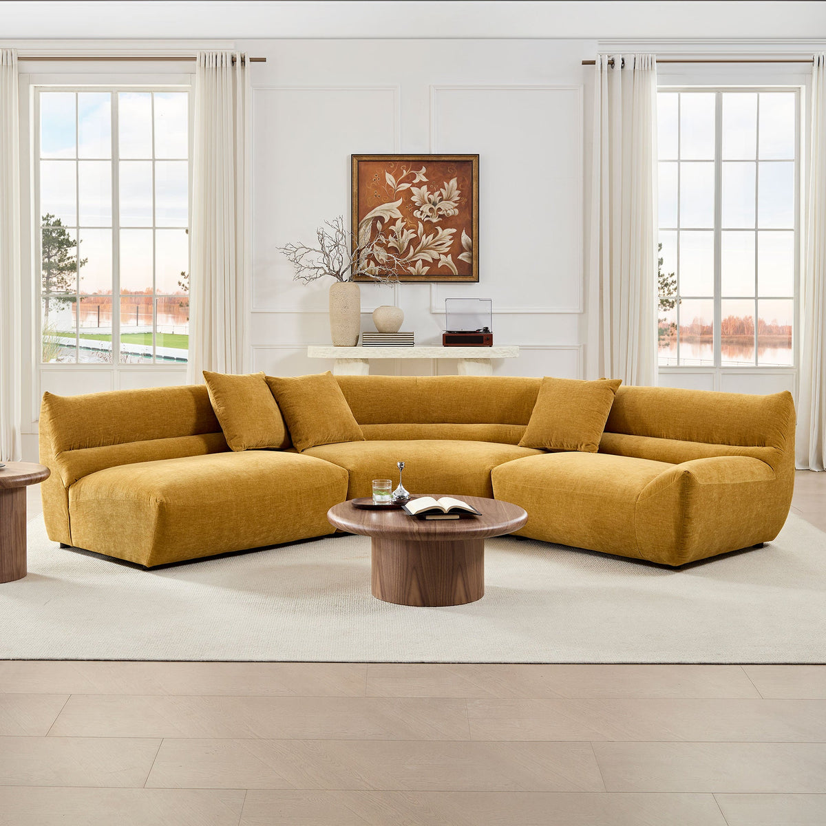 A mustard-yellow modular curved sectional sofa, placed against a white wall with windows showing a scenic view. The room is neatly decorated with a small table featuring a book and glass, and has a minimalist aesthetic.