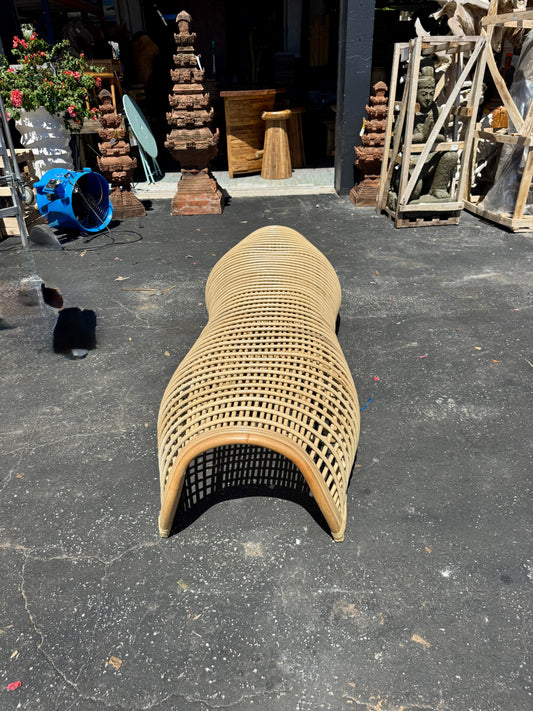 A curved woven rattan bench with a grid pattern, placed on a paved ground with a workshop background.
