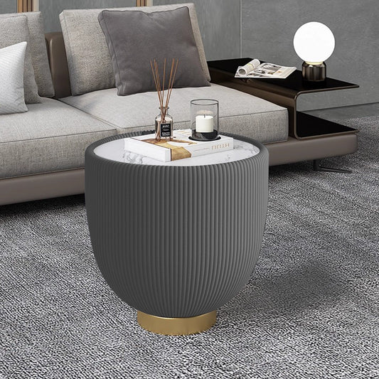 A grey ribbed side table with a marble top and gold base, displayed in a modern living room with a sofa and a smaller table in the background.