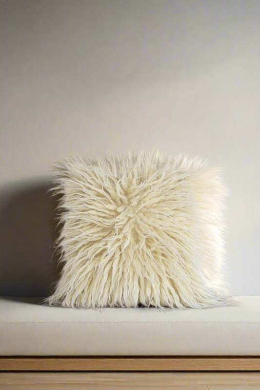 A white wool pillow with a textured, spiky fringe, placed on a light-colored sofa against a plain wall.