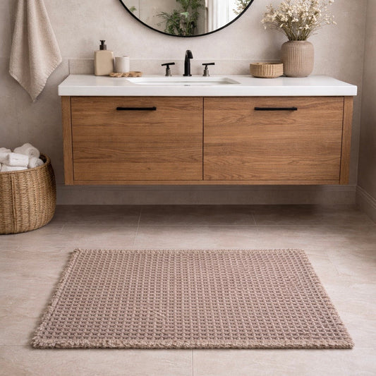 Knitted bath mat in taupe color, placed in front of a wooden vanity in a modern bathroom.