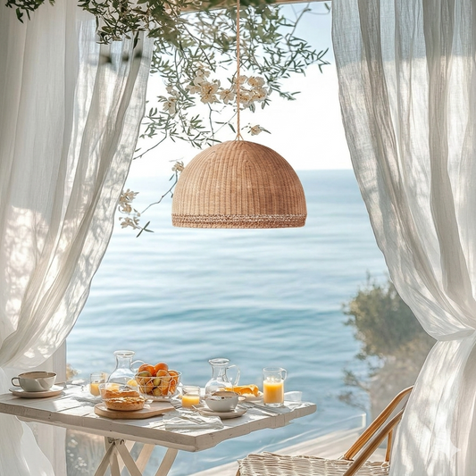 Rattan dome hanging lantern on a balcony overlooking the sea, with a table set for breakfast and white curtains, flowers, and a wooden chair.