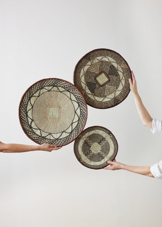 Hands holding woven baskets with lace patterns, natural fibres, brown edges, beige and dark brown, set against a plain white background.