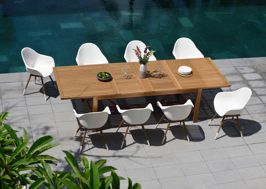 Teak rectangular table and 8 white armchairs near a swimming pool with foliage in foreground.