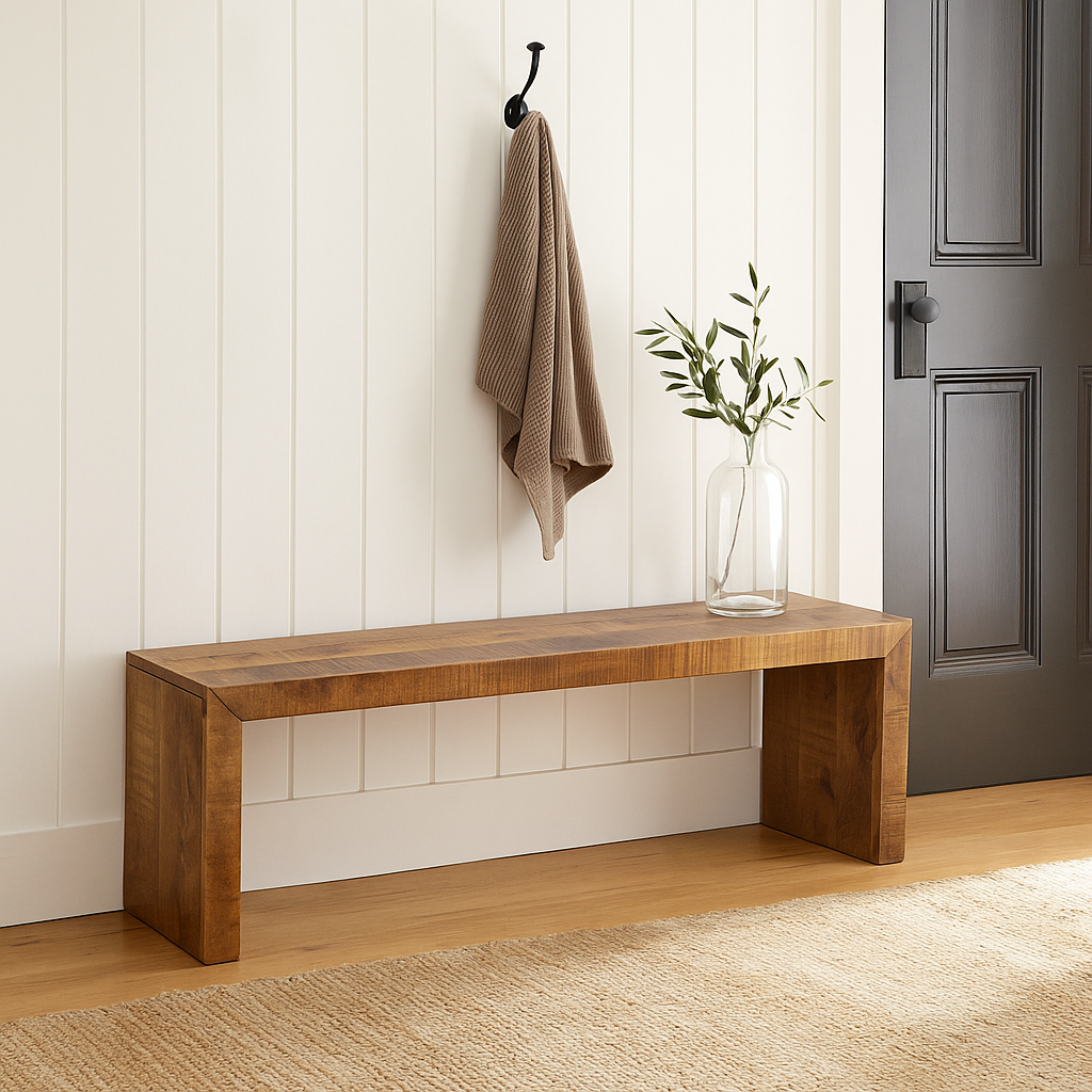 A solid mango wood bench with a light stain, resting against a paneled wall. A brown towel hangs on a hook above the bench, and a vase with greenery sits on the floor to the right.