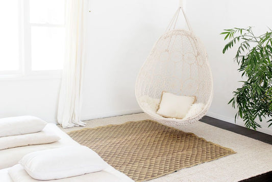 Woven hanging chair with cushions, placed on a decorative rug with white bedding nearby, in a simply decorated room.