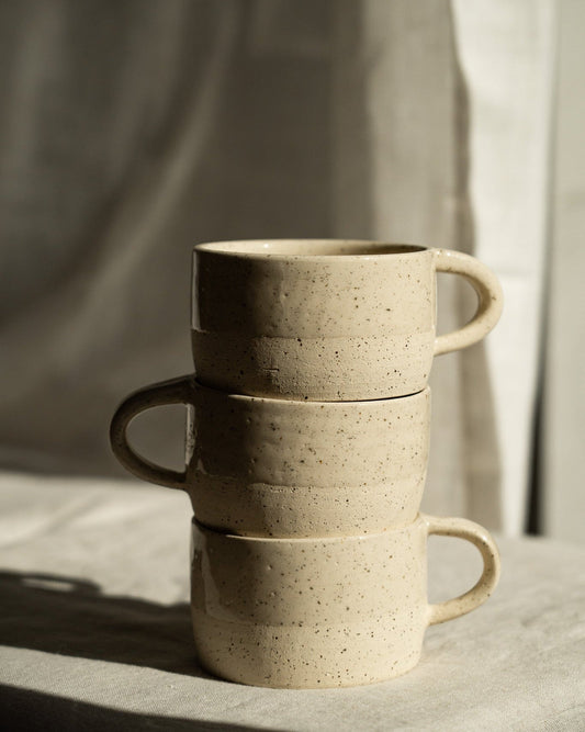 Stack of three handmade stoneware mugs with white sand speckled texture on a fabric surface in natural light