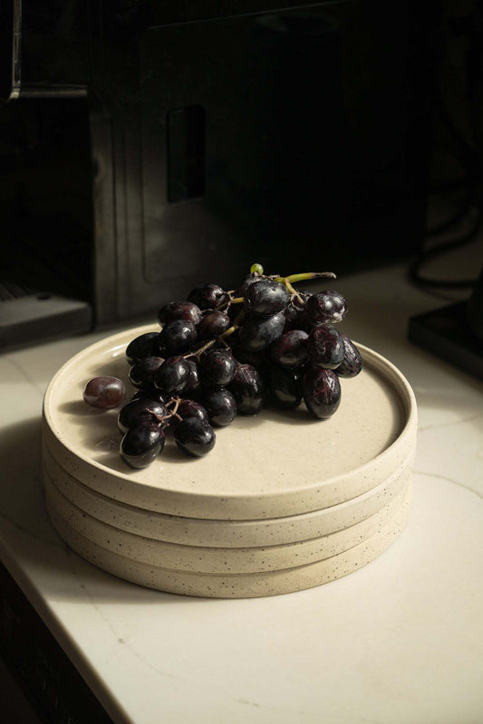 A stack of handmade stoneware pot plates with white speckled finish. On top of the stack is a bunch of black grapes.