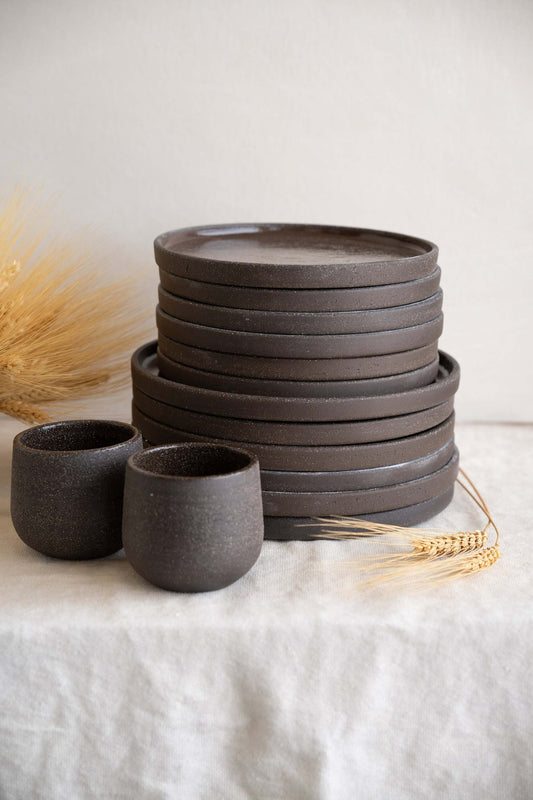 Stack of dark brown handmade stoneware dinner plates and small bowls with dried wheat as decoration on a linen cloth