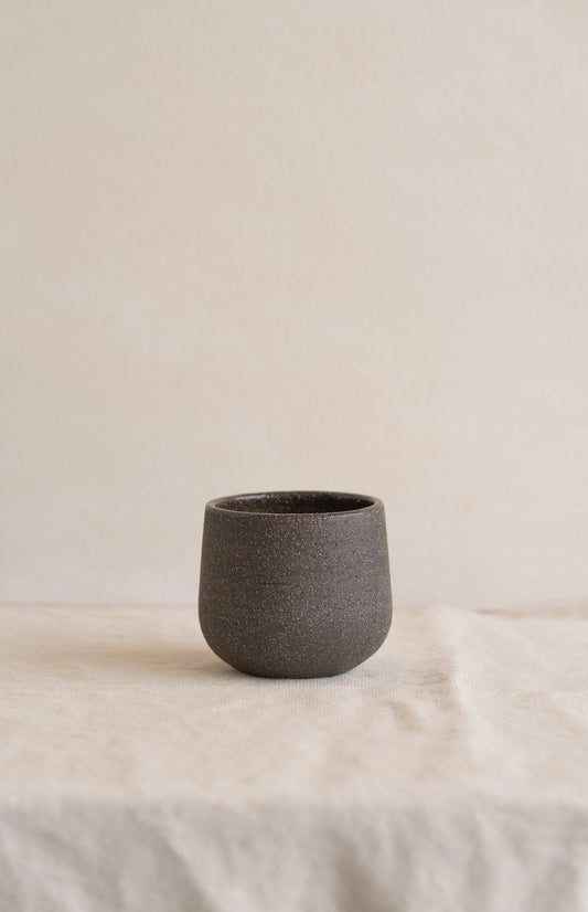 Small, dark stoneware cup with uneven texture on white fabric surface.