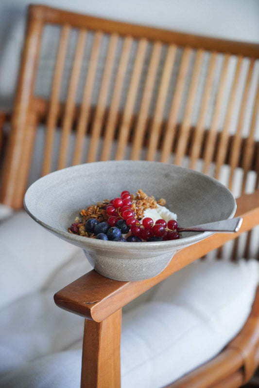 Handmade stoneware bowl shaped like a straw hat, containing granola, berries, and yogurt, placed on a wooden chair.