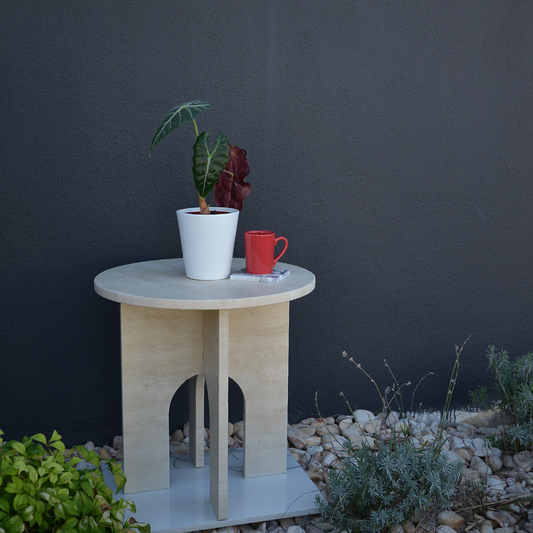 Travertine Marble Round Coffee Table with a white pot and plant, red mug, and magazine on top, situated outdoors on a white base with rocks and greenery in the background.