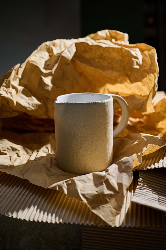 A handmade stoneware pitcher with a matte finish placed on crumpled parchment paper, possibly against a dark background.