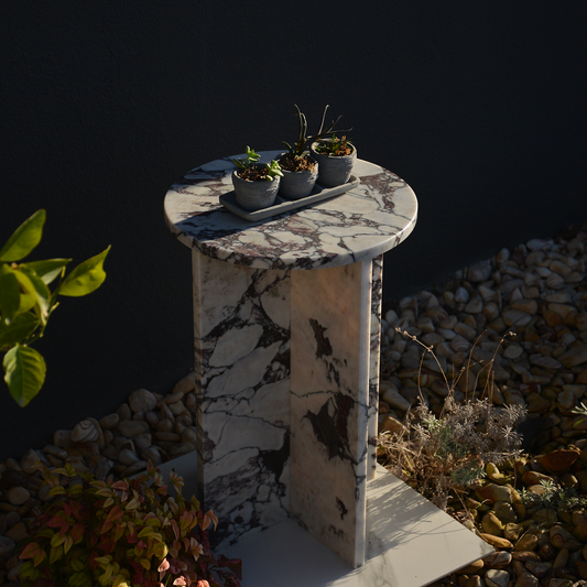 A Calacatta Viola Italian Marble Round Coffee Table with a white base and purple veining, adorned with three small potted plants on a tray, set against a dark background with rocks and a plant in the foreground