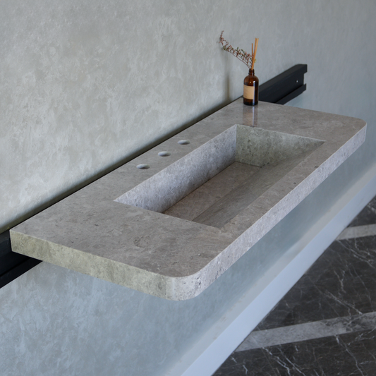 Long rectangular handcrafted sink made of marble, Tundra Grey. Sleek, modern design. Gray wall and black fixtures in the background.