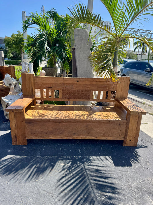 Recycled teak bench with a natural finish, placed outdoors on a paved surface with shadows of palm fronds.