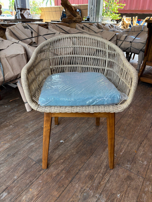 A wicker armchair with wooden legs and a blue cushion, wrapped in plastic, on a wooden floor with wrapped objects in the background.
