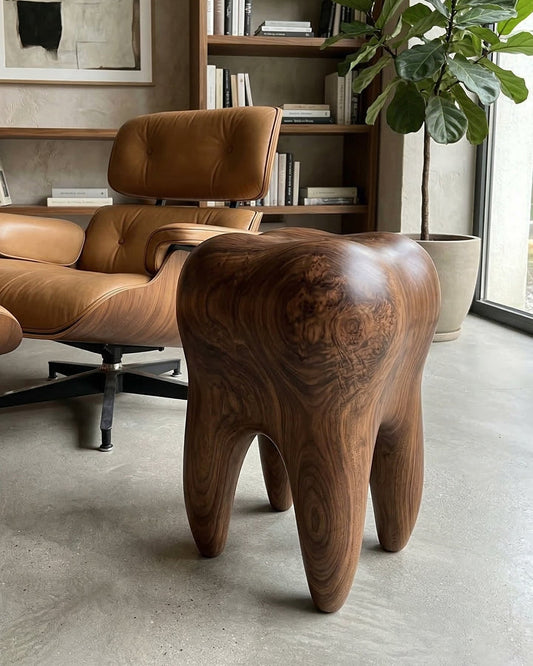 Wooden, sculpted stool resembling a tooth with wood grain and glossy finish, placed in a room with a beige armchair and bookshelf with books, natural light from window, and a potted plant.
