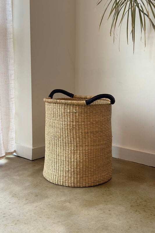 Natural woven pot basket with black handles, placed on a concrete floor in a minimalist setting.