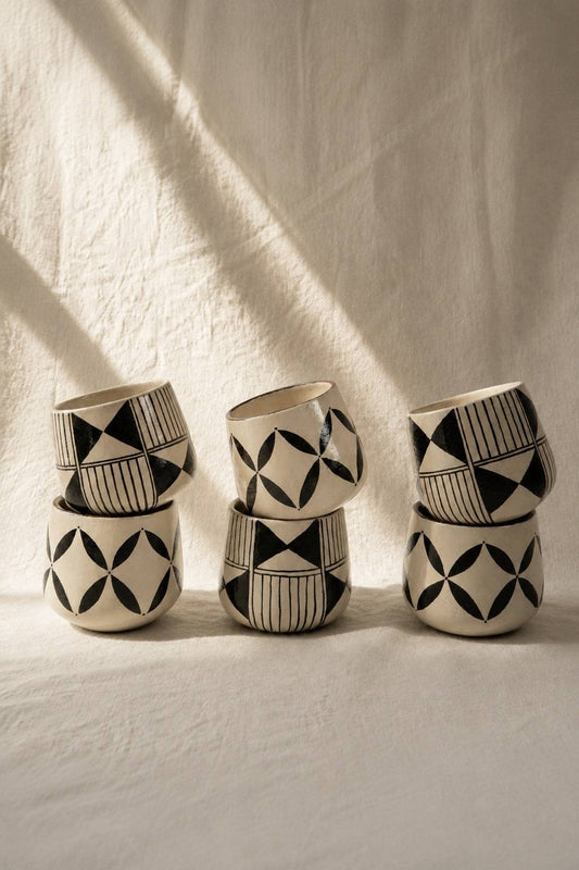 Three sets of hand-painted coffee mugs with black geometric patterns on white, stacked on a textured backdrop with soft lighting shadows.
