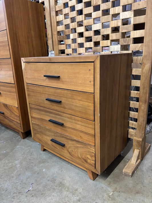 Suar 4-Drawer Dresser with natural wood finish and black metal handles in a warehouse setting