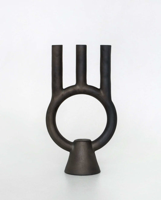 A black ceramic candle holder with three central tubes and a ring linking them, placed on a small conical base, against a white background.