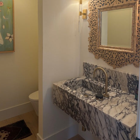 Marble sink with mixed grey and white veining against a backdrop of similar veined marble, gold faucet, wall decoration, and framed artwork. Setting appears to be a bathroom.