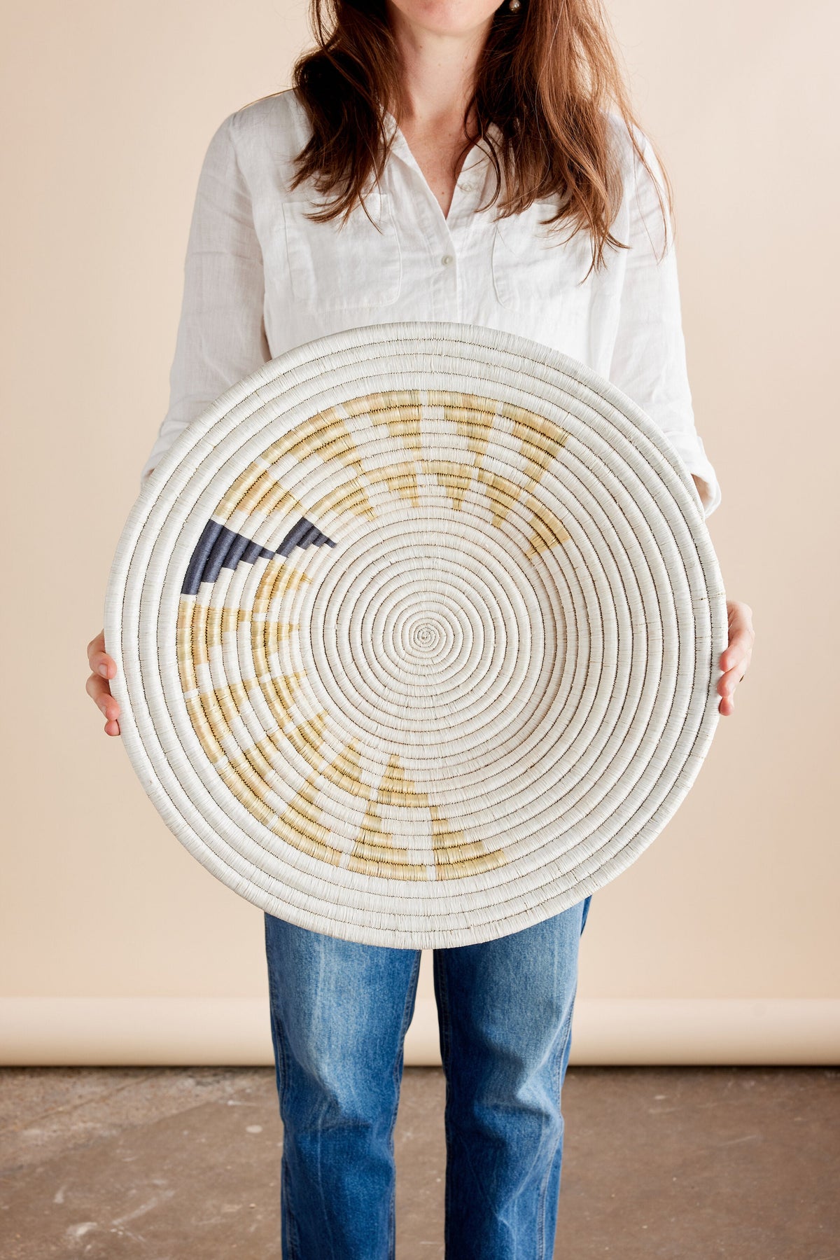 A person holding a large, circular, woven basket with a light brown and white pattern. The basket has a natural material finish and is presented against a plain, neutral backdrop.