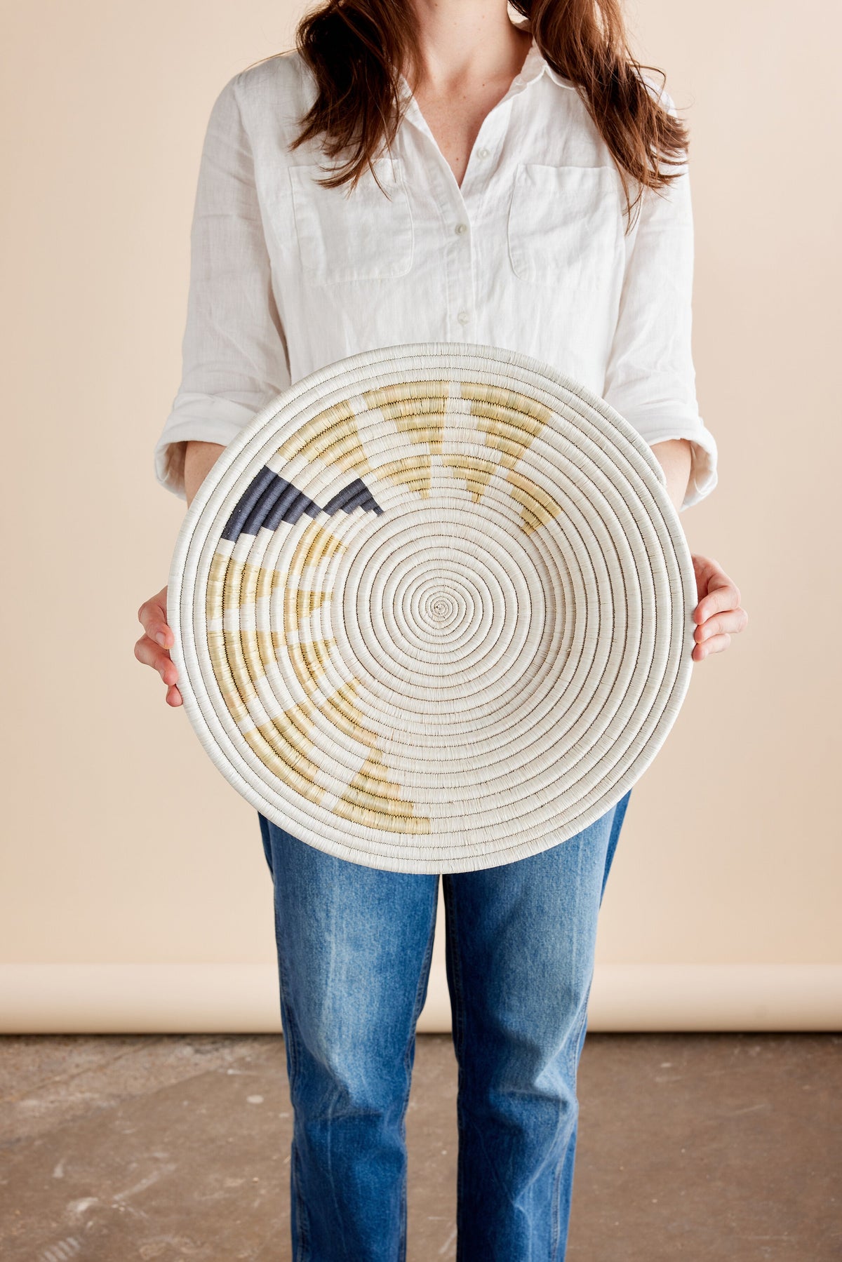 Person holding a circular woven wall basket with concentric patterns in white, beige, and navy. Neutral backdrop, indoor setting.