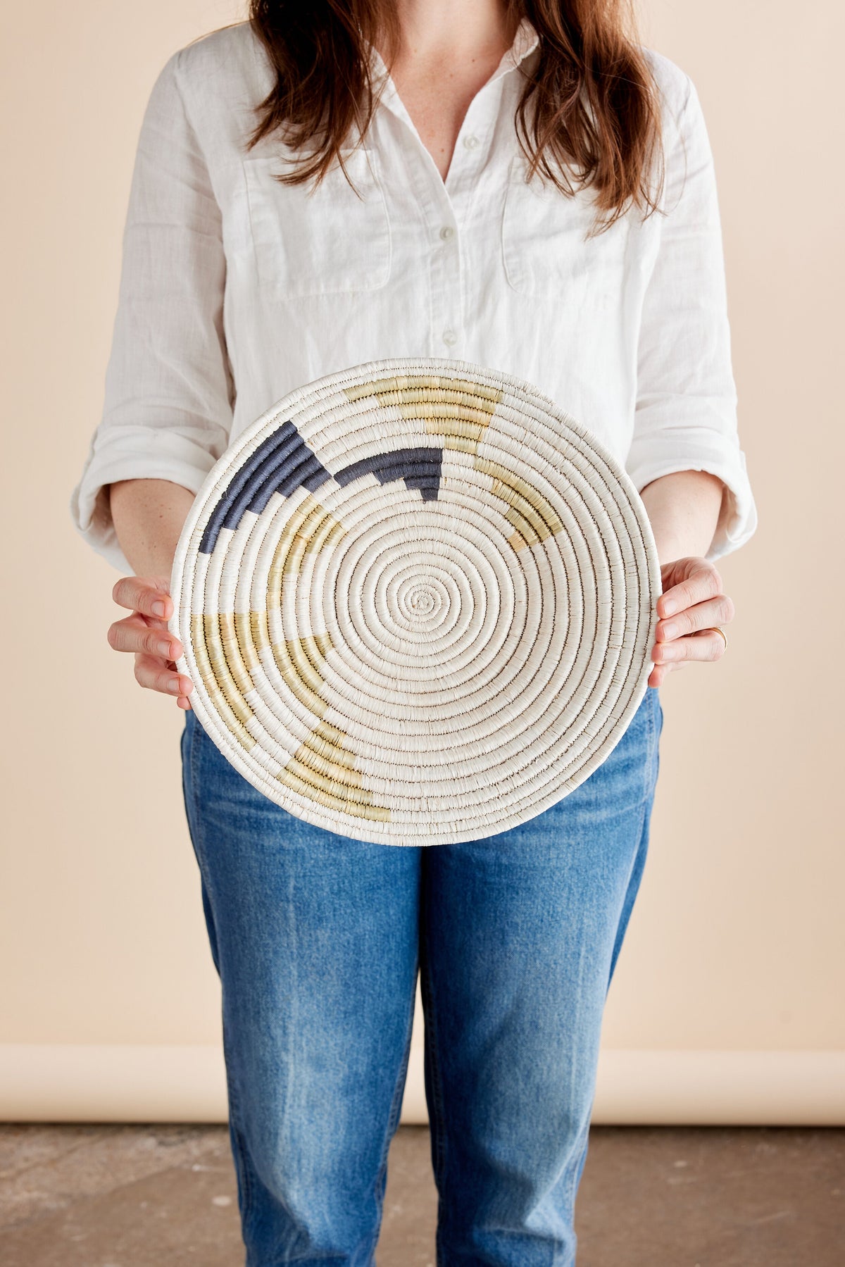 Woman holding a round wicker wall basket with a spiral pattern and a dark color accent in a minimalist setting