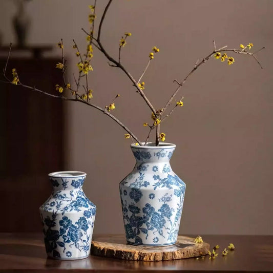 Two blue and white porcelain vases with floral designs, one taller than the other, both holding branches with small yellow flowers, displayed on a wooden base against a neutral wall.