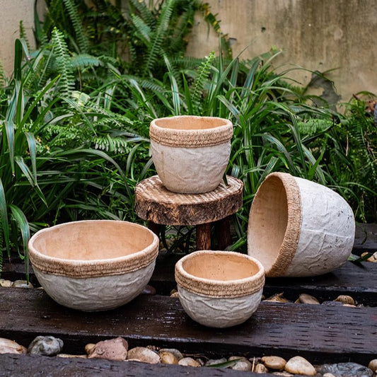 Natural ceramic planters with intricate textures and a striped edge design, placed on wooden tables outdoors, surrounded by green foliage.