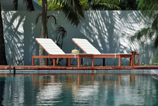 Dark wood lounge chairs with white cushions by a swimming pool surrounded by palm trees.