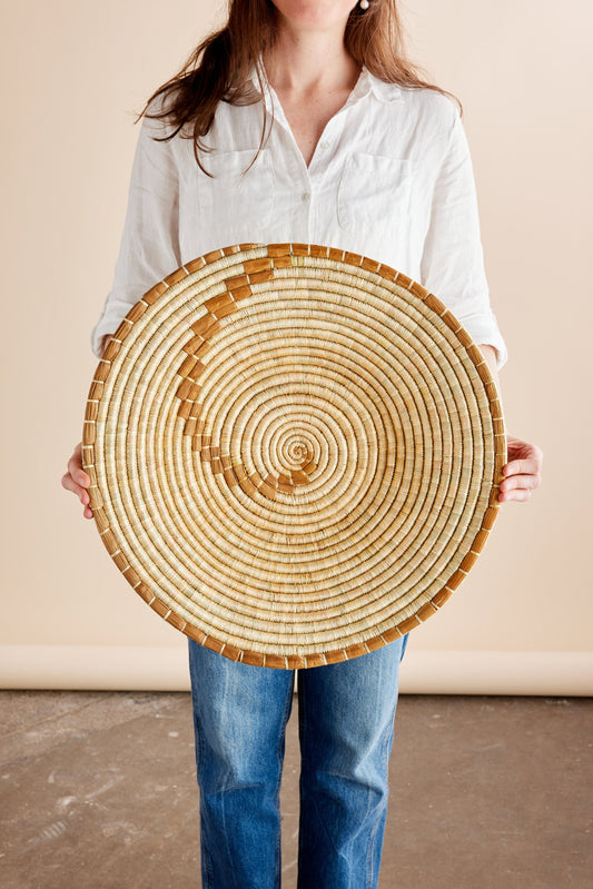 Circular rattan basket with spiral pattern held by a person in a white shirt and jeans, against a neutral background.