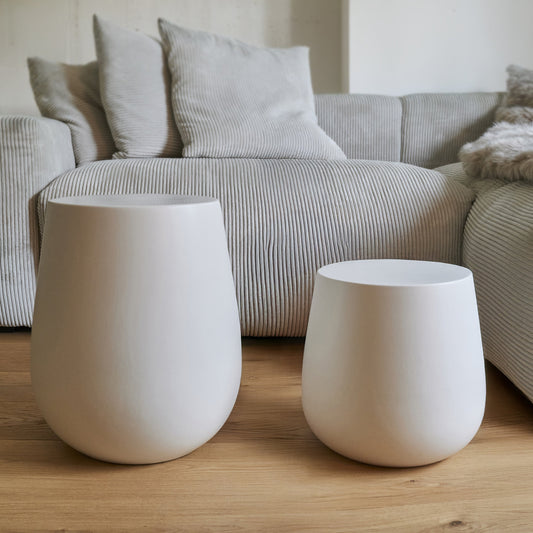 Two large, white ceramic side tables in a living room setting with a neutral sofa and cushions behind them, on wooden flooring.