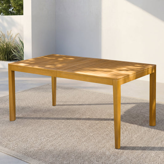 A rectangular wooden dining table with a natural finish, placed on a textured outdoor carpet with a light backdrop of white walls and some greenery.