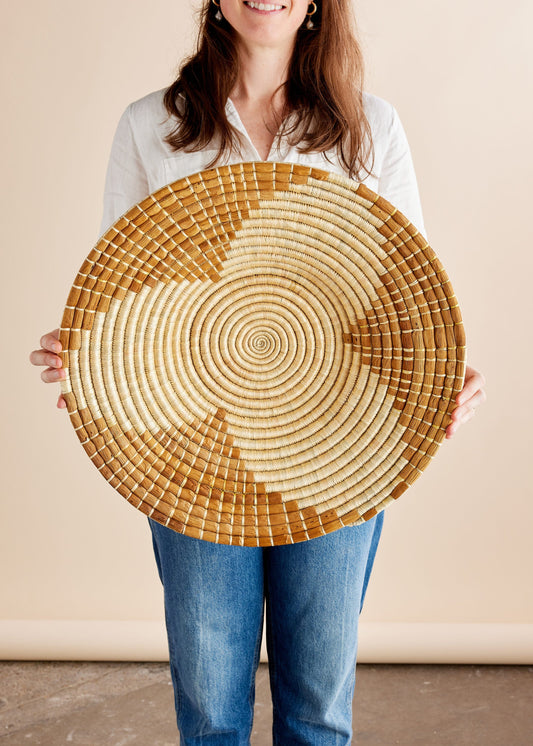 Person holding circular woven basket with spiral pattern. Material looks natural, possibly bamboo or straw. Neutral background setting.