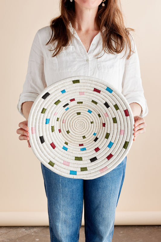 A woman holding a circular woven wall basket with a spiral pattern and multicolored accents against a beige background, wearing a white shirt and jeans.
