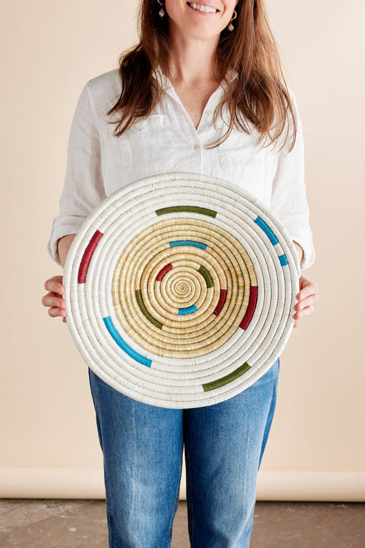 A person holding a circular woven wall basket with a spiral pattern and colored accents.