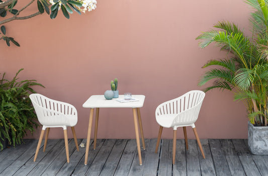 Two white Nassau Square Tables with Social Plastic® material and natural wood legs, with two matching chairs, displayed against a pink wall with plants, on a gray wooden deck.