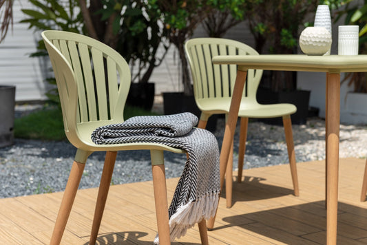Two Sage Green Nassau Side Chairs with natural wooden legs, one draped with a herringbone throw, arranged around a table with decor, outdoor setting.