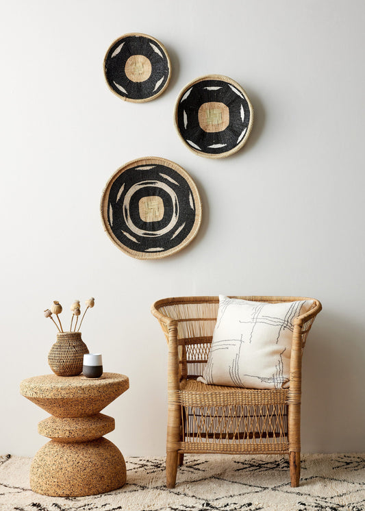 Three black and beige decorative wall baskets hanging above a wicker chair with a white cushion, next to a cork side table with a vase on it.