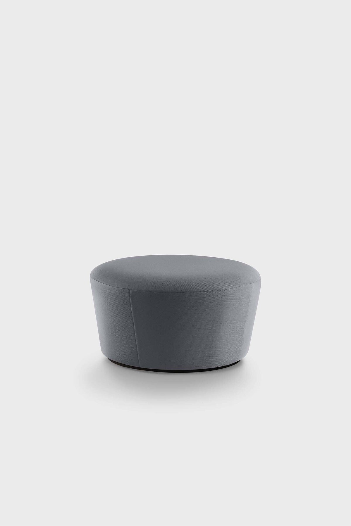 The Naïve Pouf is a round ottoman with a padded surface and a soft finish, featuring a light grey upholstery in a minimalist setting.