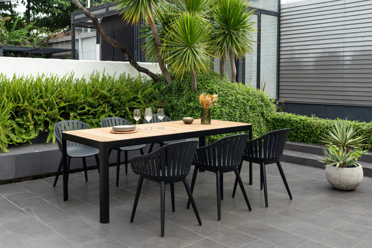 Teak dining table with black aluminum chairs on a modern patio, surrounded by greenery and hedges.